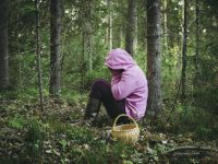 Confused person went to mushroom picking and got lost in the forest, disoriented scared and confused, northern Europe, sit under tree. Person lost in nature concept. Wearing bright colorful clothing.